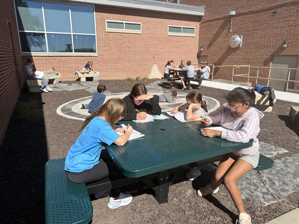 Students sitting around a green picnic table outside a brick building writing in workbooks, with additional students visible studying in the background.