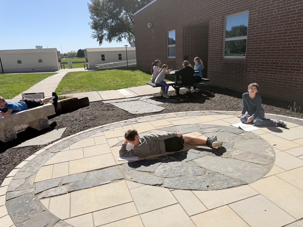 A wide view of students dispersed across an outdoor courtyard, sitting at a picnic table and lounging on stone seating while reading.
