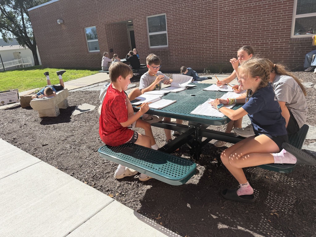 Children seated around a green metal picnic table outside doing schoolwork, with another student lying down on a stone bench nearby.