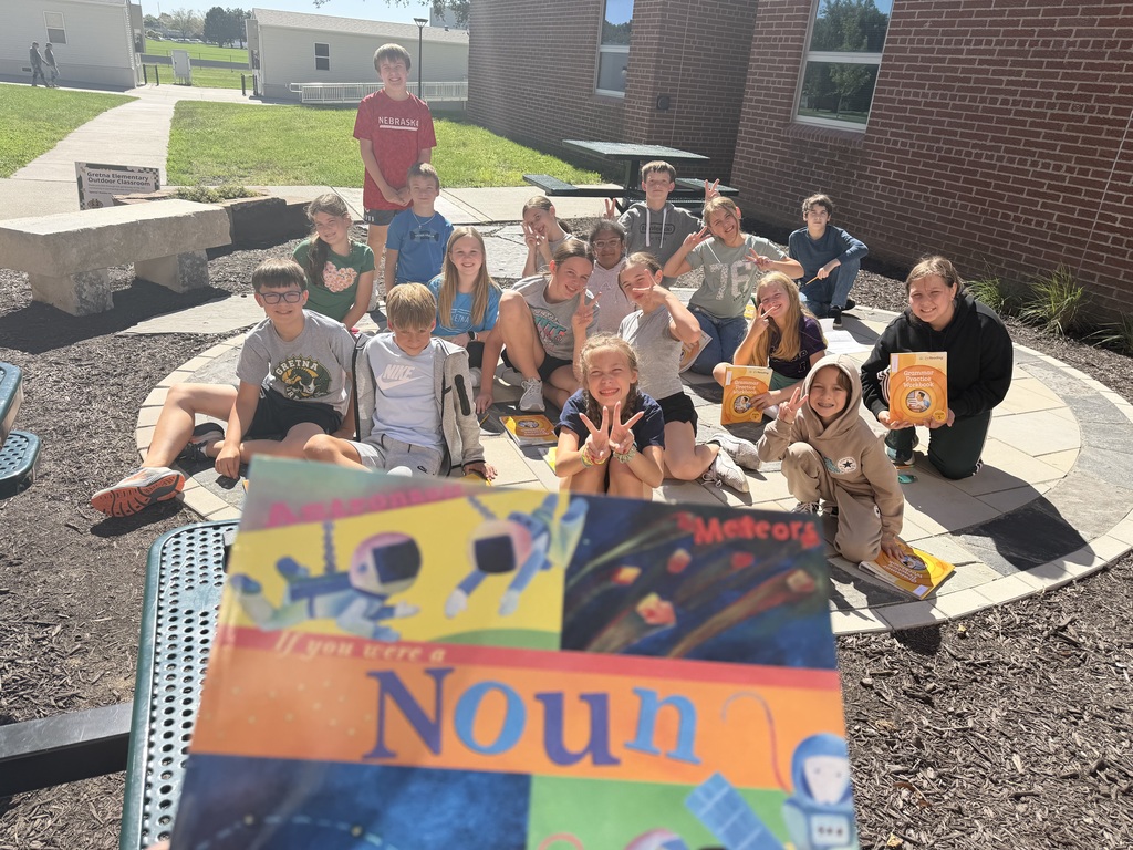 A group of elementary students gathered on a patio area posing together, with a book titled "If You Were a Noun" held up in the foreground.