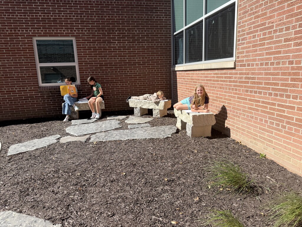 Students sitting and lying on stone benches along a brick wall outside, reading books and working on assignments.