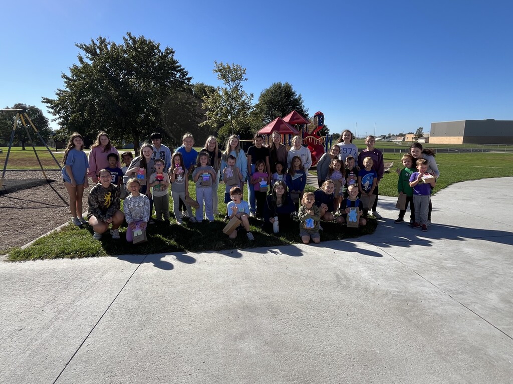 Group of students and children posing outside on a sunny day near a playground with decorated paper bags.