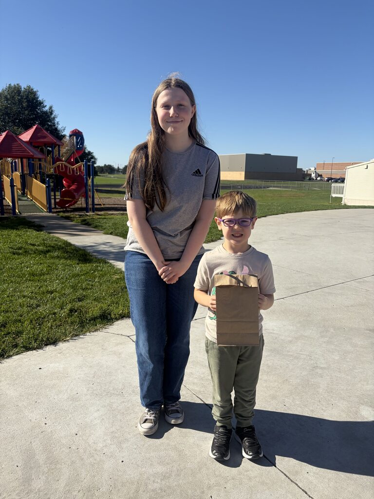 Older student standing beside a young boy wearing glasses and holding a brown paper bag outside near a playground.