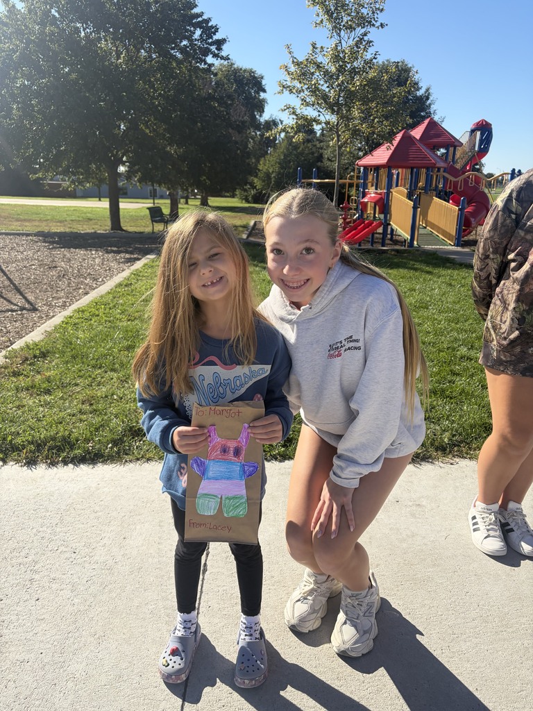 Young girl holding a colorful monster craft paper bag while standing next to a smiling older student outdoors.