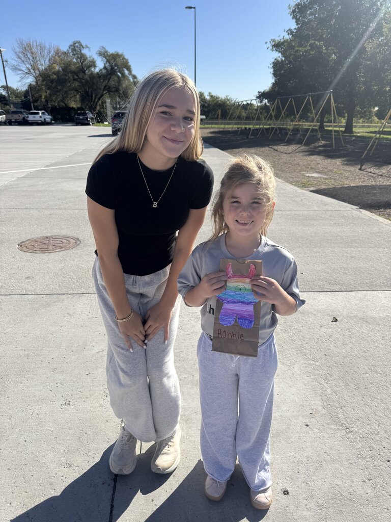 Older student leaning down next to a young girl holding a paper bag decorated with a colorful monster.