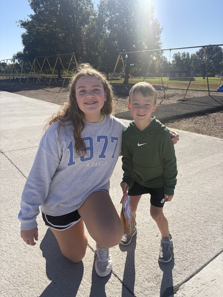 Older student crouching next to a young boy in a green hoodie holding a decorated paper bag on a concrete sidewalk.