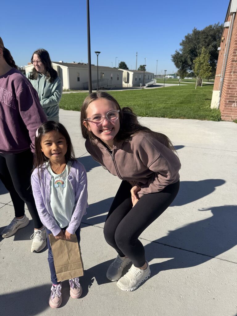 Older student leaning down next to a young girl holding a brown paper bag on a sunny outdoor walkway.