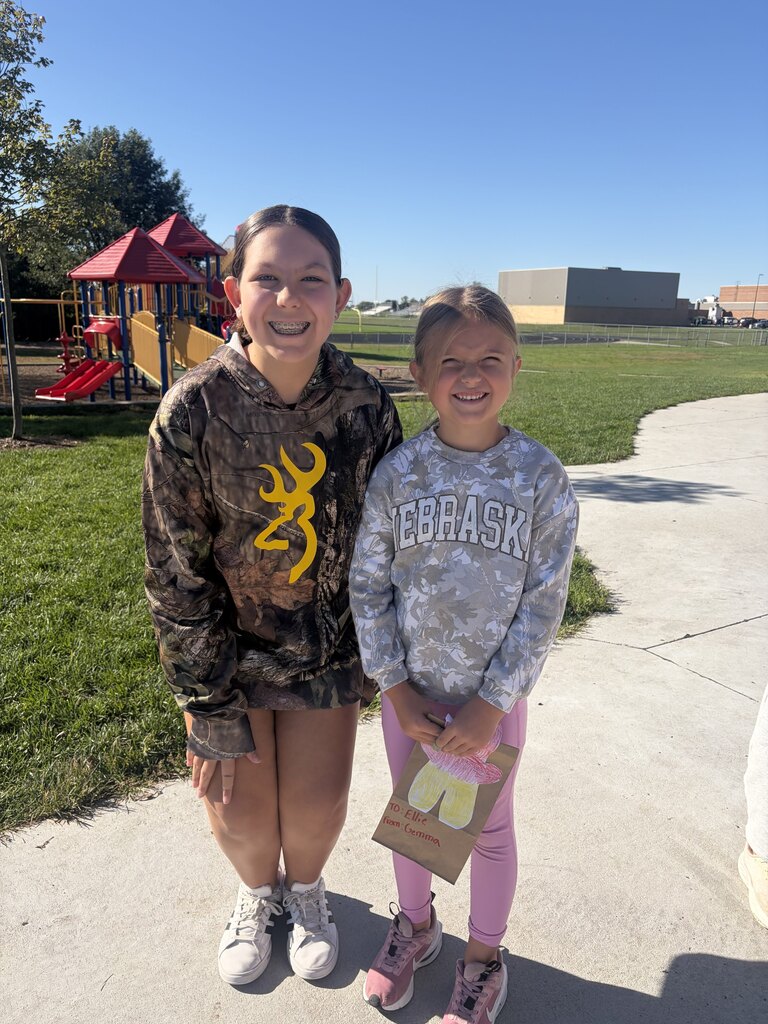 Older student in a camouflage hoodie standing with a younger student holding a decorated paper gift bag.