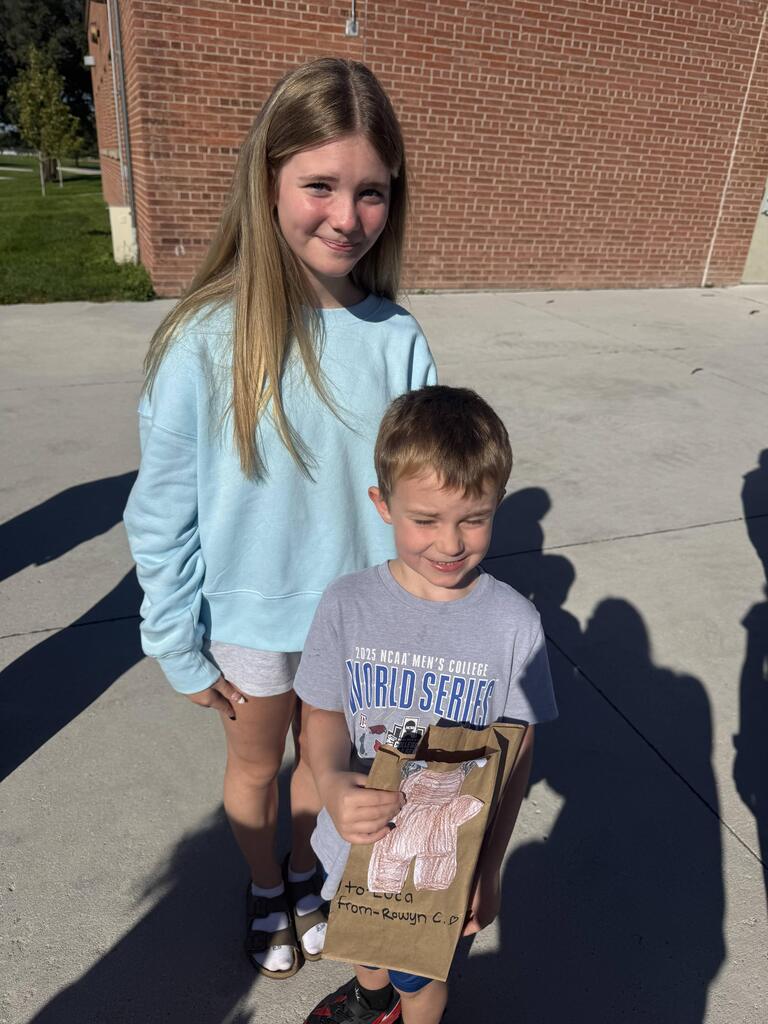 A girl and a young boy stand outside on a concrete surface with a brick wall behind them. The girl has long blonde hair and wears a light blue sweater. The boy in front wears a grey t-shirt reading "2025 NCAA MEN'S COLLEGE WORLD SERIES" and holds a brown paper bag with a drawing of a teddy bear and text reading "to Luca from-Rowyn C."