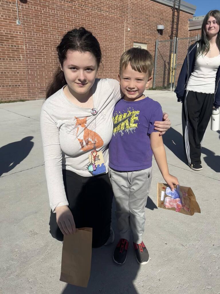 A girl in a long-sleeve cream top with a fox illustration kneels next to a young boy in a purple shirt and grey sweatpants. The boy holds a decorated brown paper bag. Another person stands in the background on the concrete paving outside the brick building.