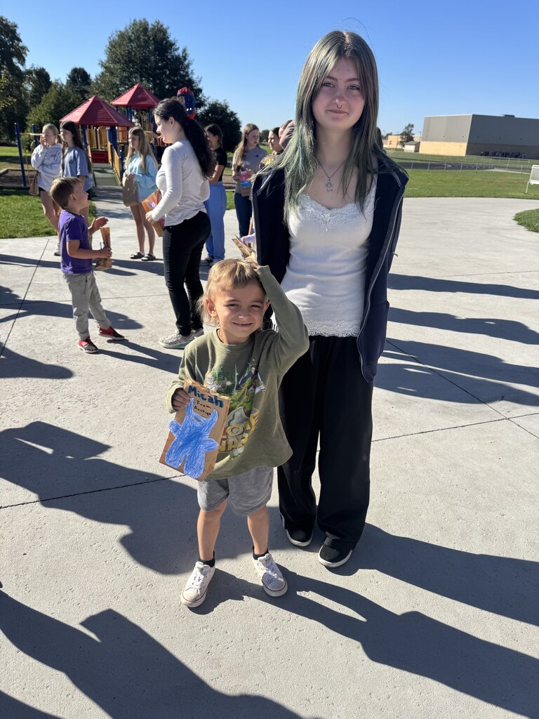A boy in a green long-sleeve shirt and grey shorts stands in front of a girl with dark hair wearing a white top and dark zip-up jacket. The boy holds a brown paper bag featuring a blue monster drawing and the name "Micah". Playground equipment and other people are visible in the background.