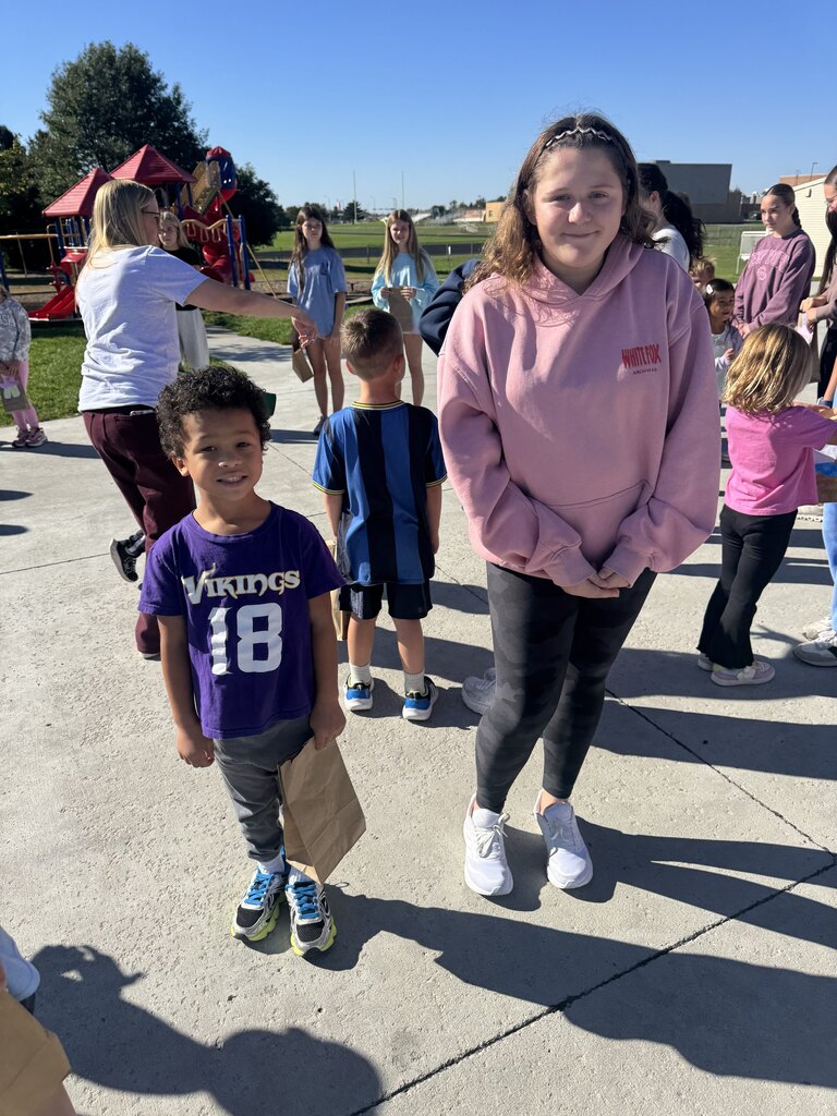 A boy wearing a purple "VIKINGS 18" t-shirt and grey pants stands next to a girl in a pink hoodie and black leggings. The boy holds a brown paper bag. Other children and adults are gathered in the sunny outdoor setting in the background.