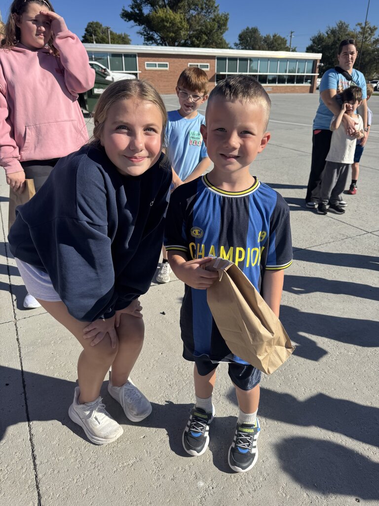 A girl in a dark navy zip-up hoodie leans down beside a boy wearing a blue and dark striped soccer jersey with "CHAMPION" on the front. The boy holds a small brown paper bag. Other children and adults are visible in the background on the concrete path outdoors.