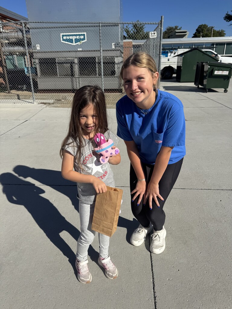 A young girl in a grey t-shirt, grey pants, and pink sneakers holds a small brown paper bag and a colorful plush toy. Beside her, a girl in a bright blue t-shirt and black leggings bends down slightly, smiling. They are outdoors on a concrete ground in front of a fenced utility area.