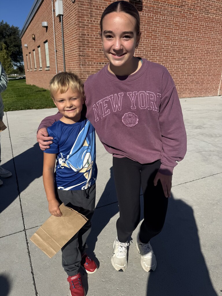 A young boy and an older girl pose together outside on a concrete walkway with a brick building in the background. The boy wears a blue graphic t-shirt featuring Sonic the Hedgehog, dark pants, and red sneakers while holding a brown paper bag. The girl wears a maroon sweatshirt with "NEW YORK" printed on it and dark leggings.