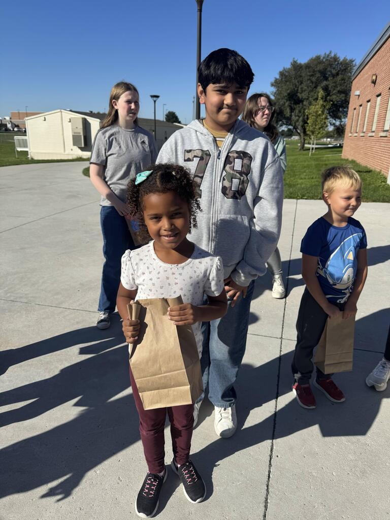 A young girl in a patterned white top and dark pants holds a brown paper bag in front of an older boy wearing a grey zip-up hoodie with "78" printed on the front. Another child holding a brown paper bag stands on the right.