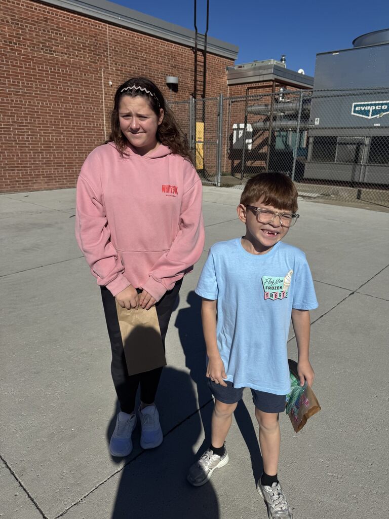 Two children stand outside on a concrete ground in front of a brick building. The girl on the left wears a light pink long-sleeve hoodie, dark pants, and white shoes, holding a small brown paper bag. The boy on the right wears a light blue t-shirt with a graphic, dark shorts, glasses, and grey sneakers, also holding a small brown paper bag.