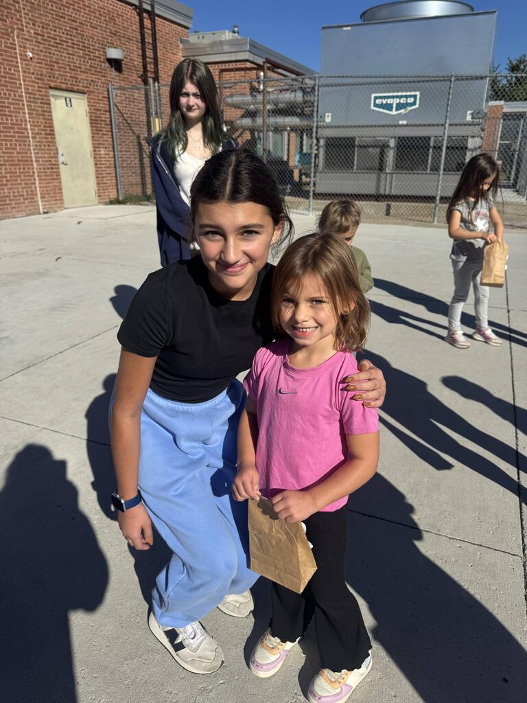 A girl in a black short-sleeve shirt and light blue sweatpants crouches down next to a younger girl wearing a bright pink t-shirt and dark pants. The younger girl holds a brown paper bag. In the background, another girl stands near a brick wall and industrial equipment, while another child stands nearby holding a brown bag.