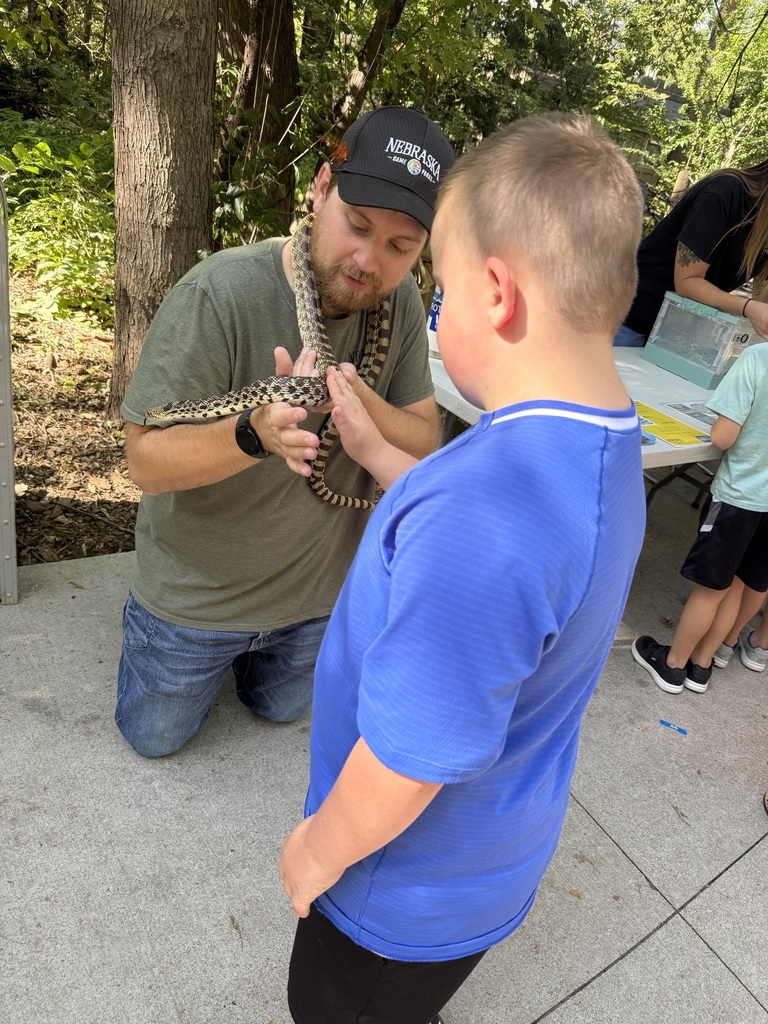 A boy in a blue shirt touches a snake wrapped around the neck of an instructor wearing a black Nebraska Game and Parks cap. They are outdoors next to a demonstration table.