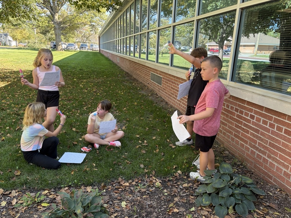 Five children gather outside on the grass near a brick school building, holding papers and eating popsicles while engaging in an activity together.