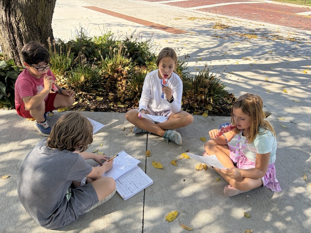 Four elementary students sit cross-legged on a concrete sidewalk under the shade of a tree, eating popsicles while reading and writing on papers and notebooks.