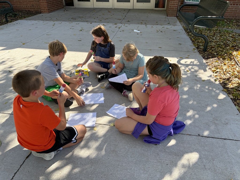 Five elementary students sit in a circle on a concrete outdoor patio while eating colorful popsicles and reviewing handwritten papers spread around them.
