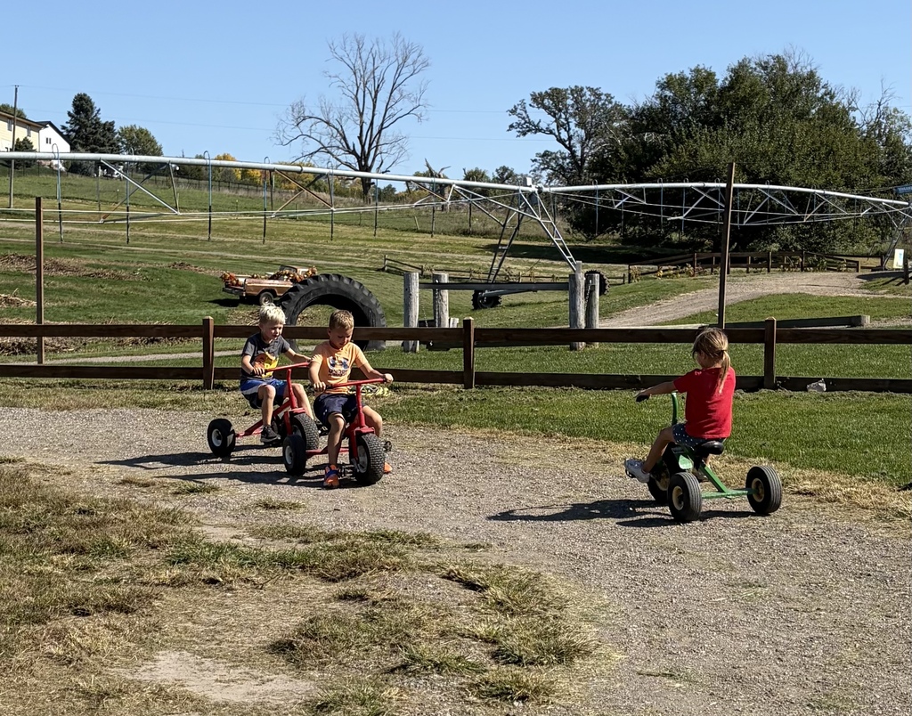 Three children riding oversized pedal tricycles along a dirt track outdoors with farm machinery and fields in the background.