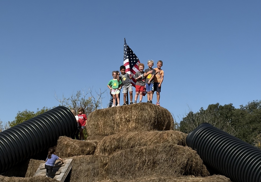 Group of school children standing atop a large mound of stacked hay bales with an American flag flying behind them on a clear blue day.
