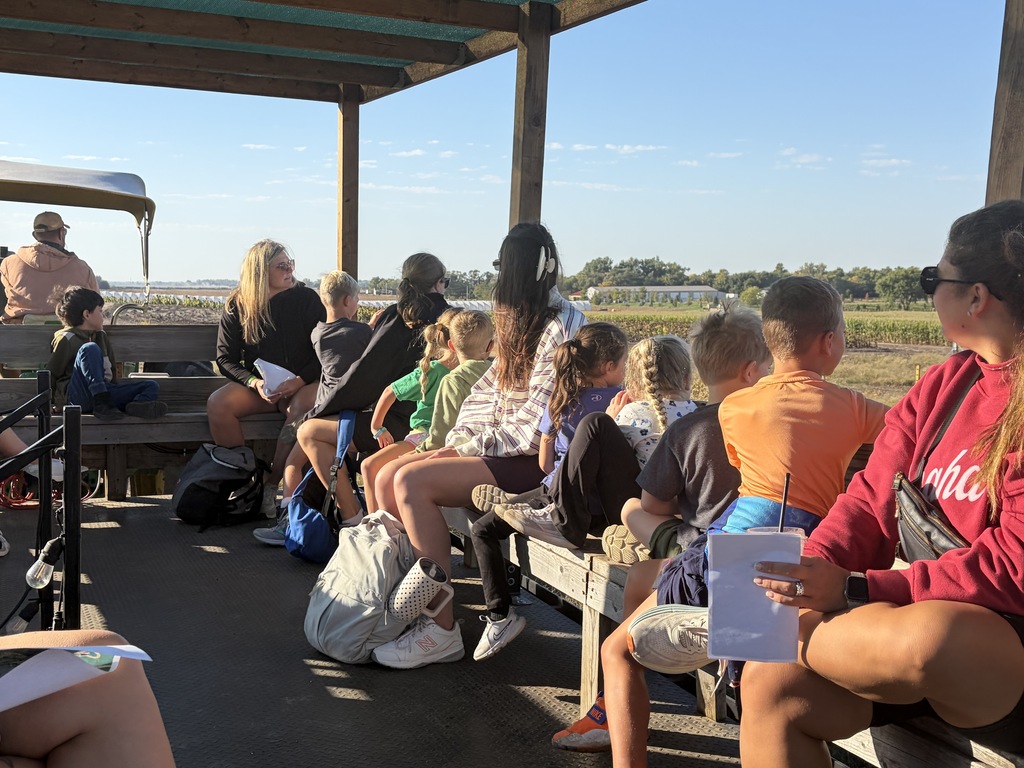 Group of adults and children sitting together on wooden benches aboard a covered wagon ride, looking out over a open field on a clear, sunny day.