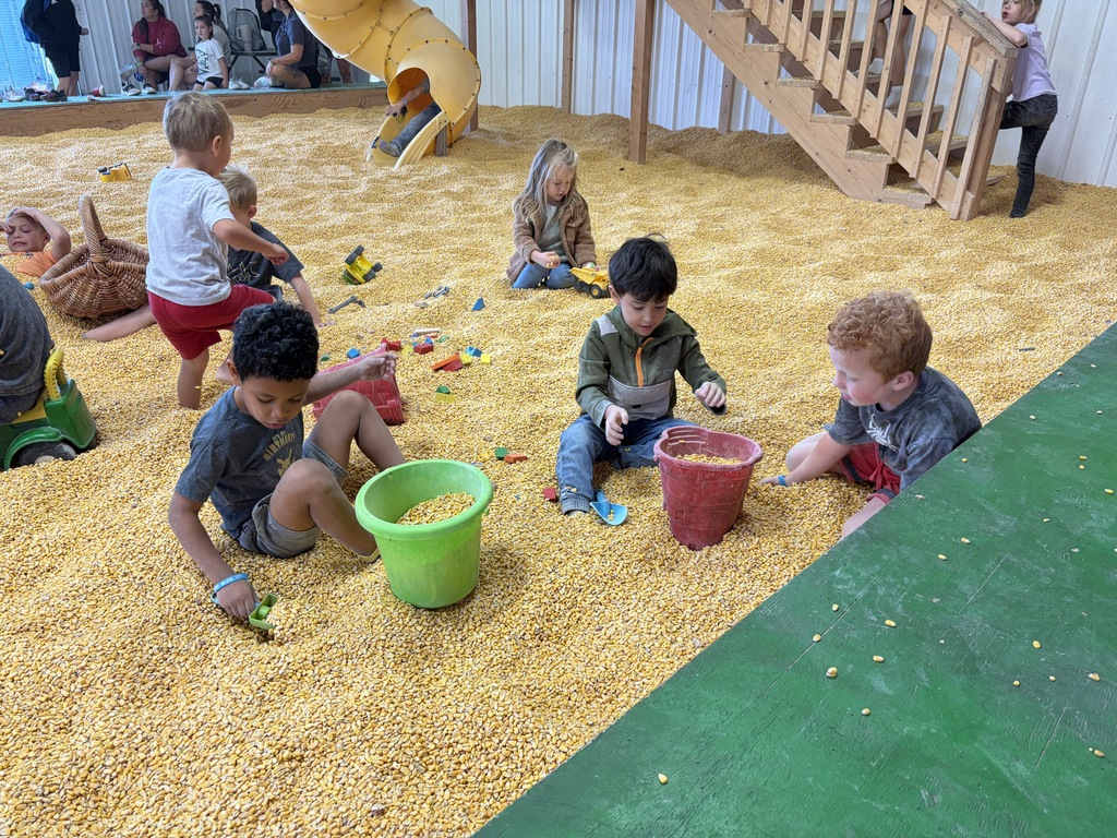 Children playing inside a large indoor corn pit filled with dried corn kernels, using buckets, shovels, and toy trucks near a wooden slide and staircase.
