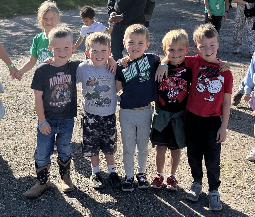 Five young boys standing side-by-side with their arms around each other on a dirt path outdoors, smiling for a group photo.