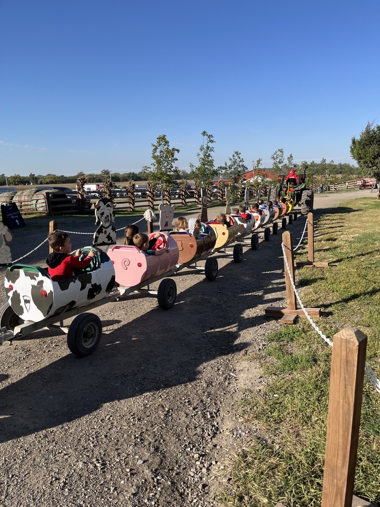 Line of barrel train cars styled like farm animals carrying children down a gravel path, pulled by a tractor in the distance.