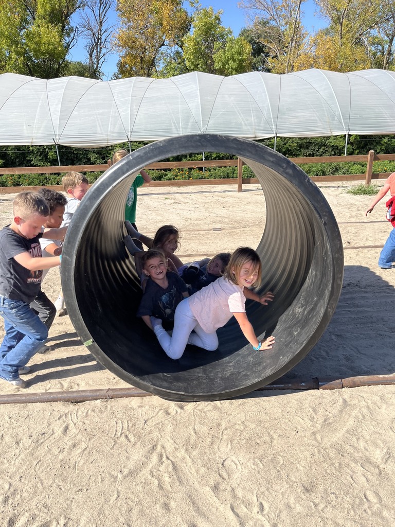 Group of young children playing and crawling inside a large black plastic drainage pipe on a sandy play area outdoors.