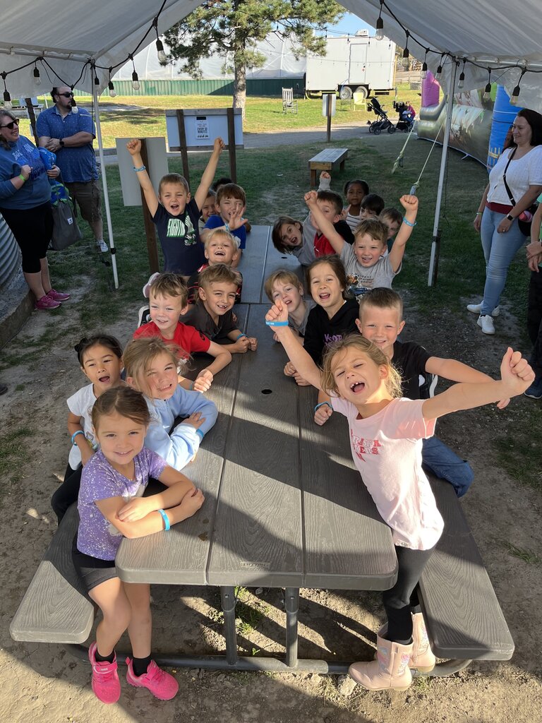 Group of young students sitting around a long outdoor picnic table under a tent canopy, smiling and raising their hands for the camera.