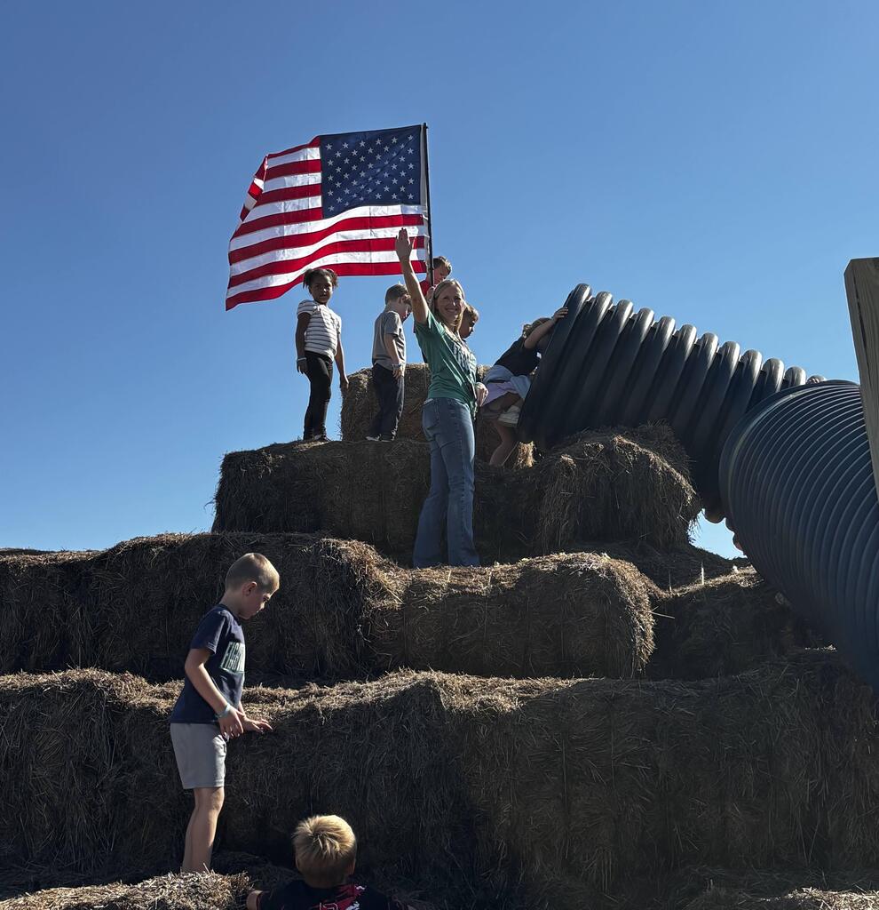 Teacher waving while standing atop a large mound of stacked hay bales next to students and an American flag under a clear blue sky.