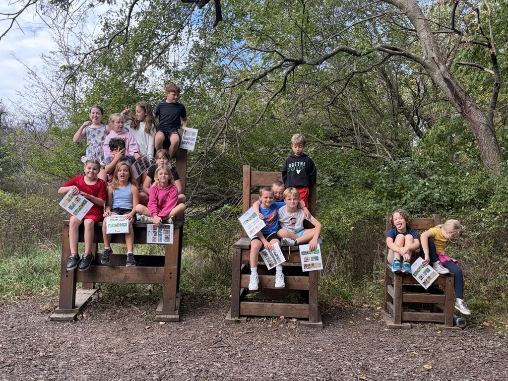 Group of young elementary students holding activity sheets while posing together on three oversized wooden chairs outdoors surrounded by trees.