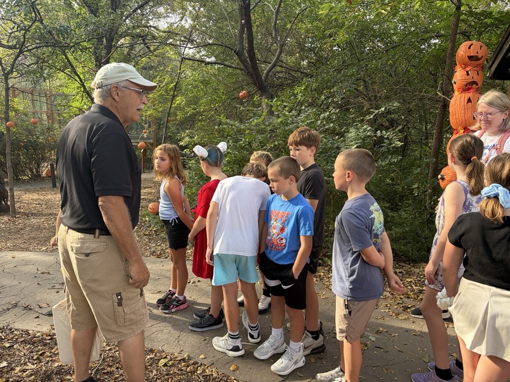 An adult guide wearing a cap and cargo shorts addressing a group of young students gathered along a paved outdoor path decorated with pumpkin figures.