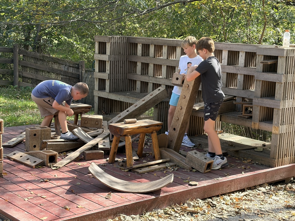 Three boys playing with large wooden building blocks and wooden beams on a red wooden platform outdoors.