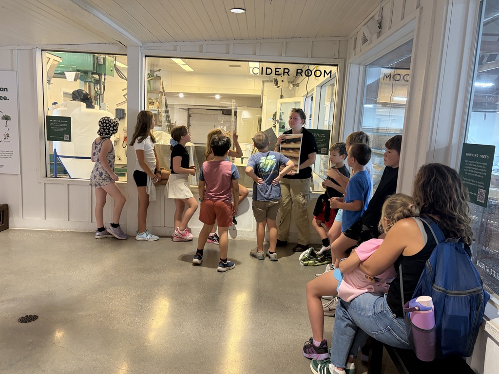 Group of students gathered inside a building, looking through glass windows into a room labeled "CIDER ROOM" while an instructor holds up an educational poster.