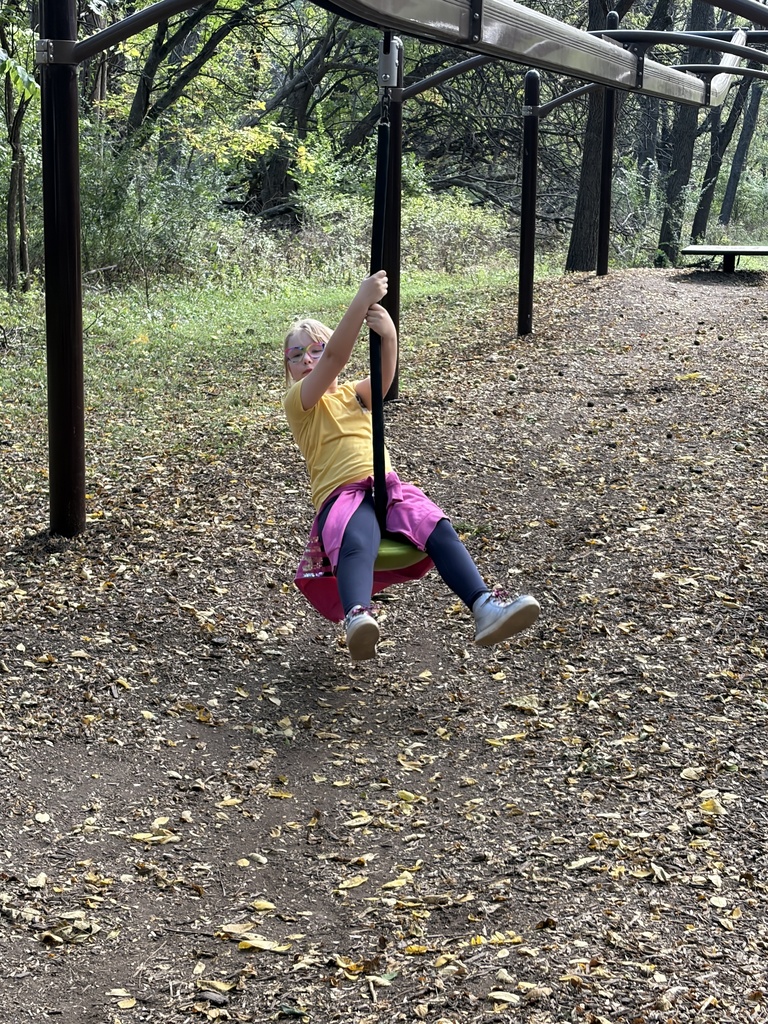 A young girl in a yellow shirt and pink jacket riding a zipline seat through a wooded playground area.