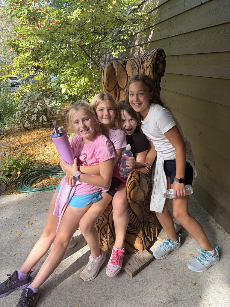 Four young girls sitting together in a large carved wooden chair on a porch, holding water bottles and smiling for a close-up photo.