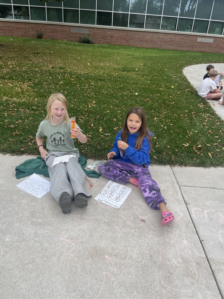 Two elementary school girls smiling while sitting cross-legged on a concrete walkway outdoors, holding test tubes and reviewing papers next to chalk drawings.