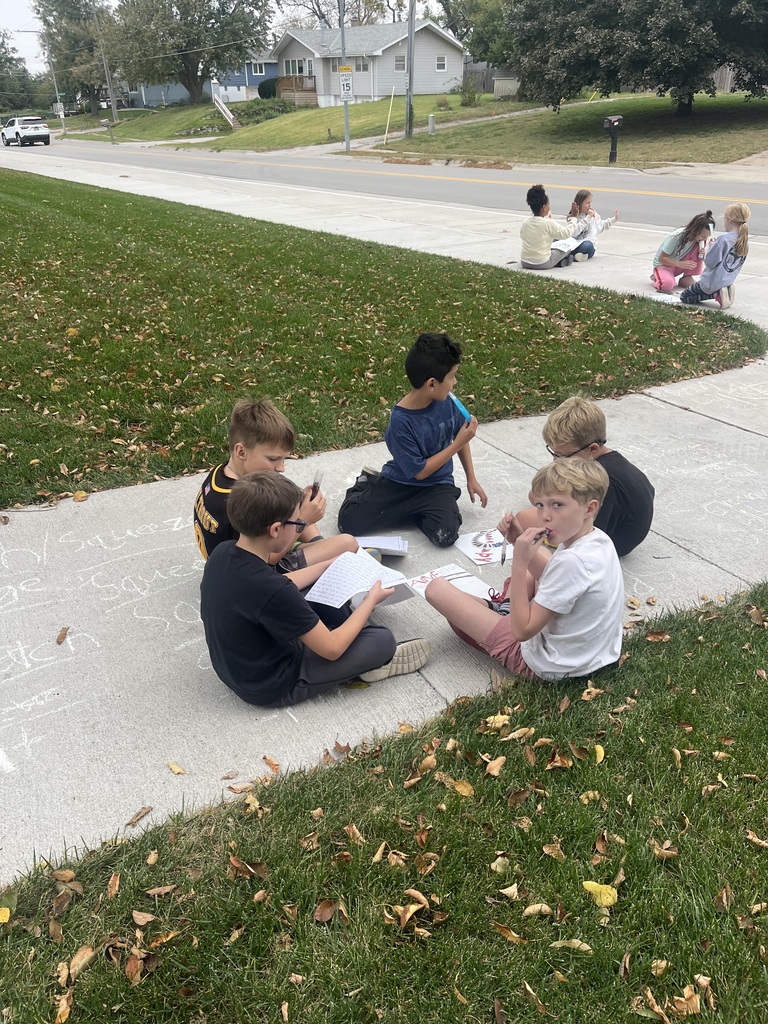 A group of young boys sitting in a circle on a sidewalk near a lawn, working together with worksheets and holding small items during an outdoor activity.