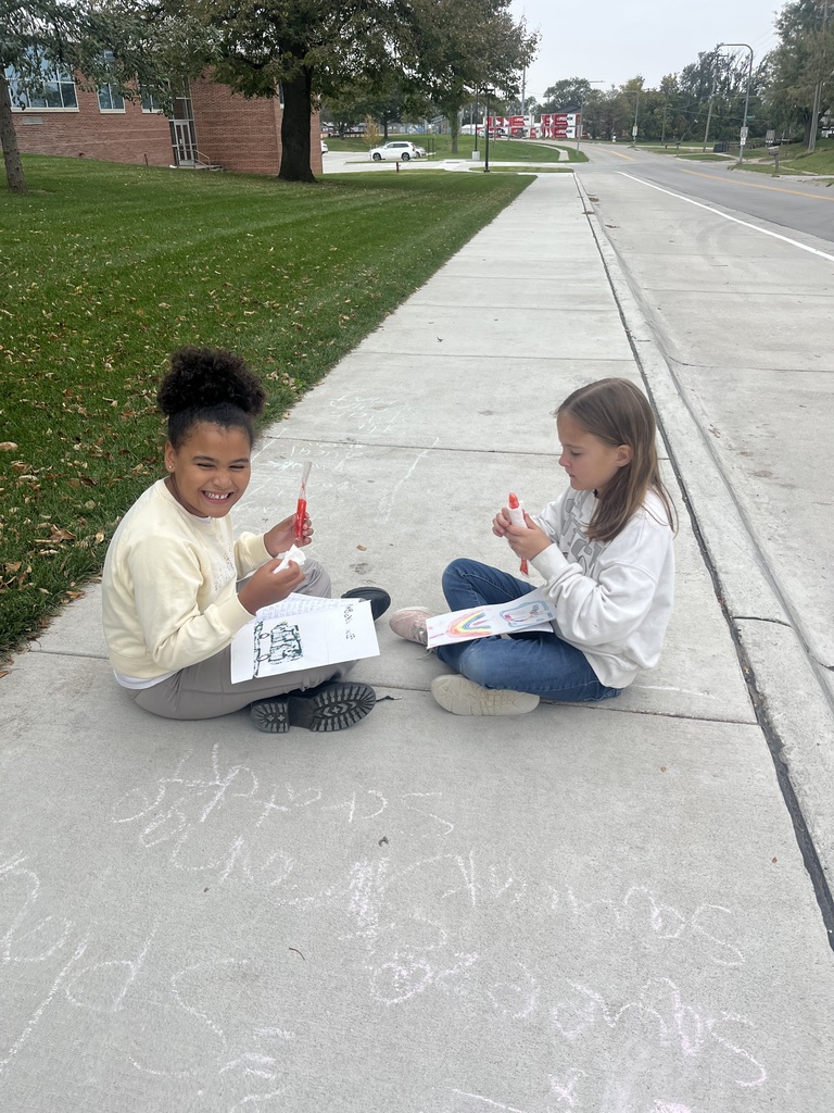 Two young girls sitting on a sidewalk outdoors, holding markers and colored sheets while smiling and completing an activity together.