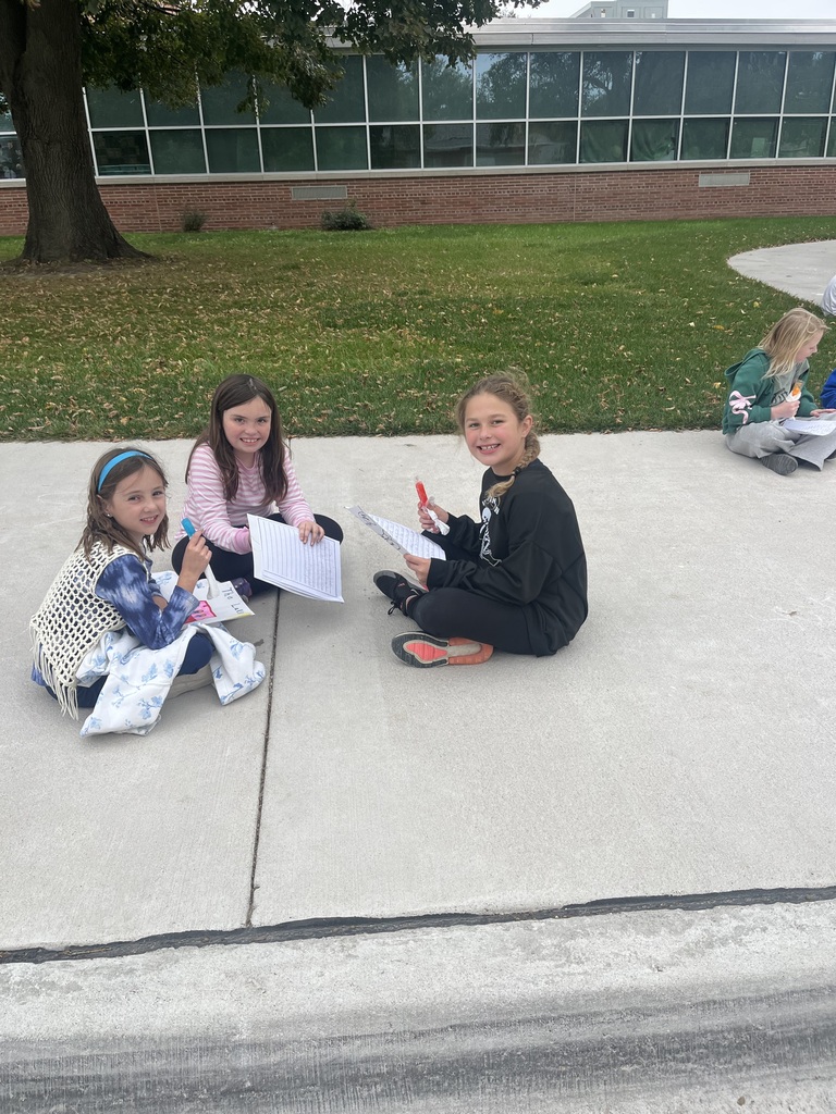 Three girls sitting cross-legged on a concrete walkway in front of a brick building, smiling while holding papers and science materials.