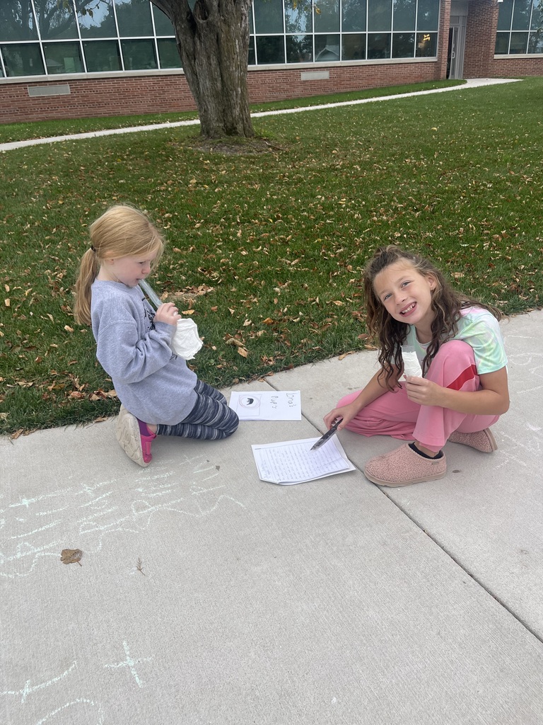 Two elementary school girls kneeling on a concrete path outside near a brick school building, holding papers and science supplies while working together.