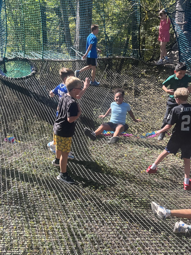 Group of children playing and climbing on a large suspended outdoor cargo net mesh play area surrounded by trees.