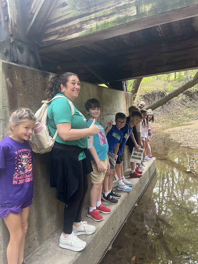 An adult chaperone and a group of young students posing along a concrete ledge beneath a wooden bridge next to a calm stream.