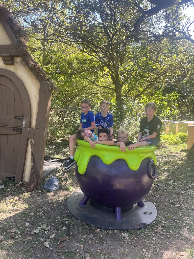 Six young boys posing for a photo inside and around a giant purple and green decorative cauldron prop outside near a small wooden hut.