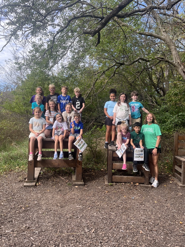A large class of elementary school students and their teacher sitting and standing together on giant wooden chairs in a wooded park area.